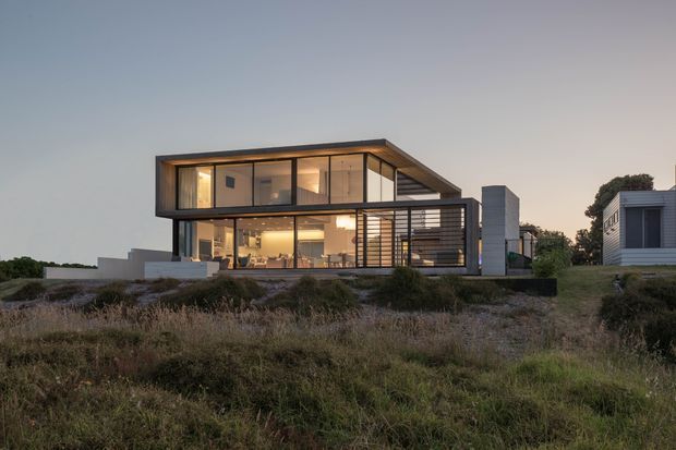 Twilight exterior photograph of a contemporary two-storey house set within a coastal-looking landscaped site. The home has a rectilinear flat-roof form, extensive floor-to-ceiling glazing, dark slender framing, projecting upper-level volumes, and a covered outdoor terrace with horizontal screening. Warm interior lighting is visible through the glass, while a smaller detached or neighbouring modern structure appears to the right.