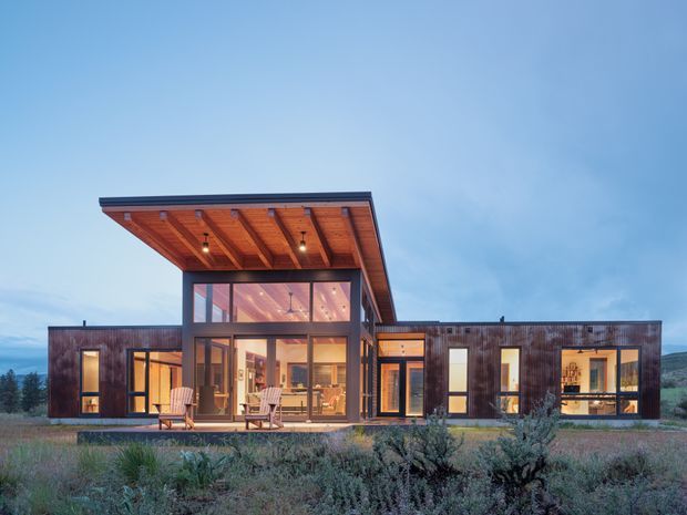 A modern single-storey residence viewed from the landscaped exterior at dusk. The home has a long, low horizontal composition with dark timber cladding, extensive floor-to-ceiling glazing, and a prominent raised central volume beneath a broad asymmetrical sloping roof. Warm interior lighting reveals living spaces through the windows, while the covered entry or outdoor area features an exposed timber soffit and integrated downlights. Low native planting surrounds the house, with hills visible in the distance.