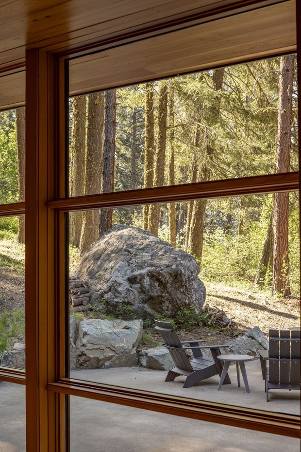 View from inside a wood-lined room through large timber-framed windows, looking onto a forested rocky site and a concrete outdoor terrace with dark lounge chairs and a small table. The glazing and warm timber framing dominate the image, creating a strong connection between the interior and the natural landscape.