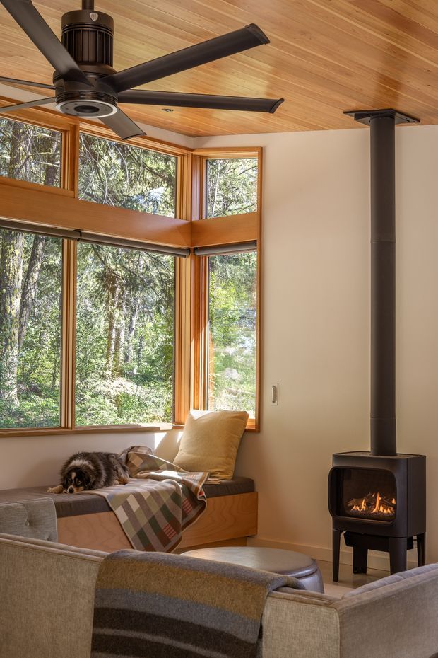 A warm, compact living room with expansive timber-framed corner windows overlooking dense woodland. The room features a timber-lined ceiling, a large dark ceiling fan, a built-in window seat with cushions, upholstered seating, a throw blanket, and a freestanding black wood-burning stove with visible fire. The space has a cozy, natural cabin-like atmosphere with abundant daylight.