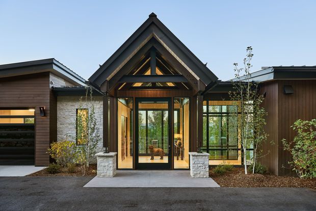 Front exterior view of a contemporary single-storey residence, centred on a prominent gabled entry pavilion with dark timber framing, glazed double doors, and clerestory glazing. Flanking wings combine horizontal timber cladding, pale masonry, and dark metal roof elements. A paved approach, low planting, and stone entry piers complete the architectural composition.