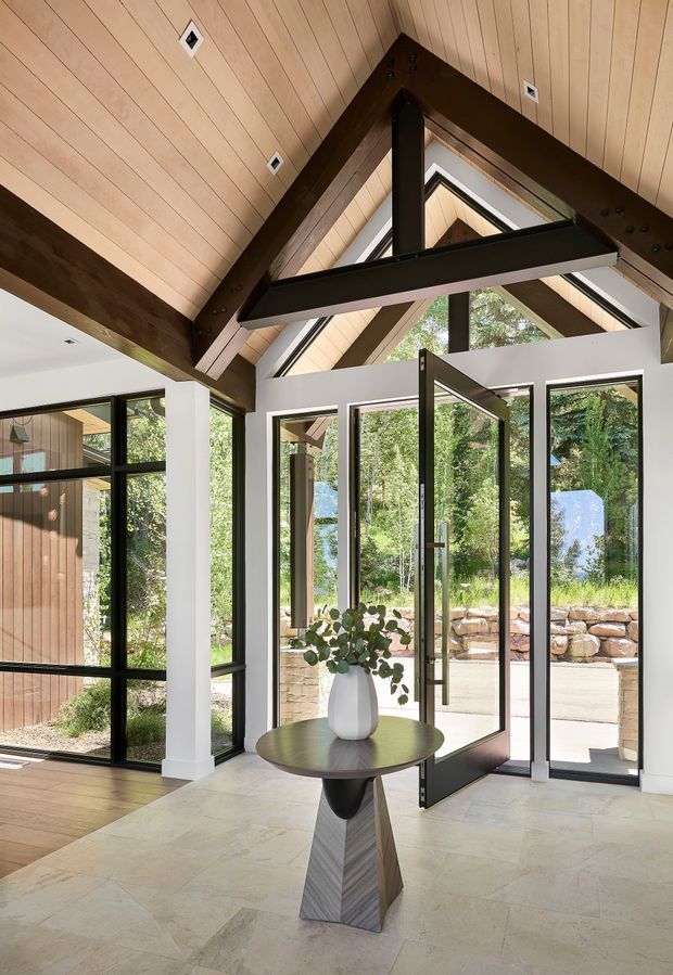A bright, double-height residential entry hall with a dramatic exposed timber gable roof and dark structural beams. Full-height black-framed glazing and a glazed entrance door open to a landscaped exterior. A small round console table with a sculptural pedestal and vase sits in the centre, while pale stone flooring and white walls create a refined, airy interior.