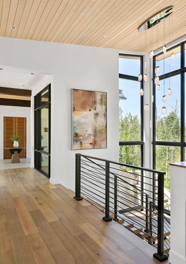 A bright, contemporary residential landing overlooking a stairwell. The space features wide-plank timber flooring, a warm timber-lined ceiling with recessed downlights, large black-framed floor-to-ceiling windows, and a black metal stair balustrade with diagonal cable detailing. A sculptural cluster of pendant lights hangs in the double-height stair void, while an abstract artwork and timber entry door add visual interest.