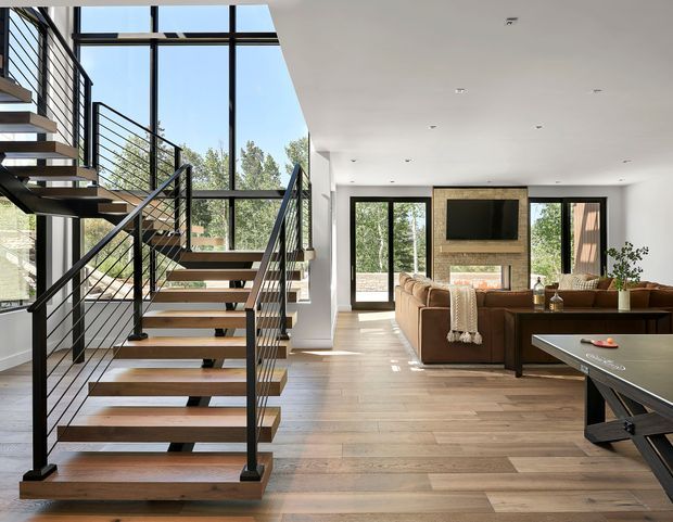 Bright contemporary open-plan living room with a floating timber staircase and black metal cable railing beside expansive floor-to-ceiling glazing. The room features wide-plank timber flooring, a brown sectional sofa, a stone or brick fireplace surround with wall-mounted television, recessed ceiling lights, and views of trees outside. The stair void and glazing create a spacious, airy interior.