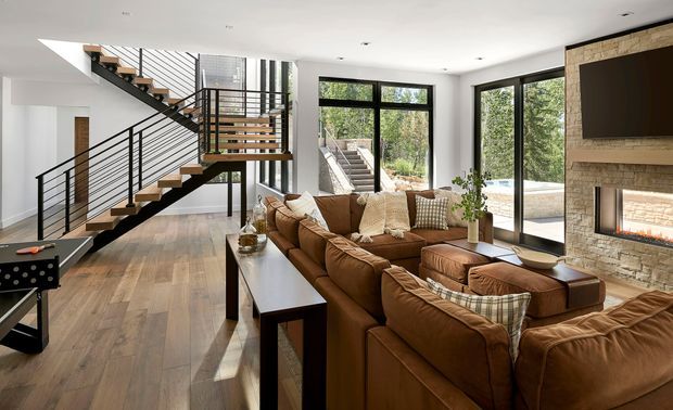 A bright, open-plan contemporary living room with a large brown leather sectional arranged around a stone fireplace and wall-mounted television. A dramatic open-riser timber staircase with black metal railings forms a prominent architectural feature, while full-height black-framed glazing opens views toward greenery and an outdoor terrace. Warm timber flooring and natural stone create a refined, inviting interior.