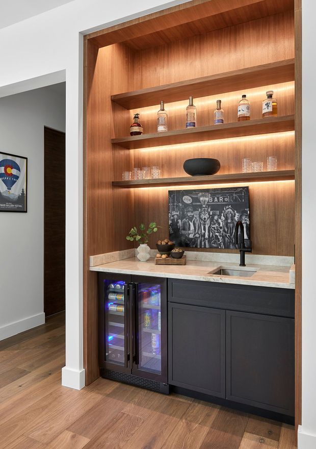 A built-in home bar set within a recessed timber-lined alcove. The bar features warm LED-lit open shelving displaying bottles and glassware, dark lower cabinetry, a pale stone benchtop with an integrated sink and matte black tap, and under-counter beverage fridges. Timber flooring and a partially visible adjacent hallway complete the contemporary interior.