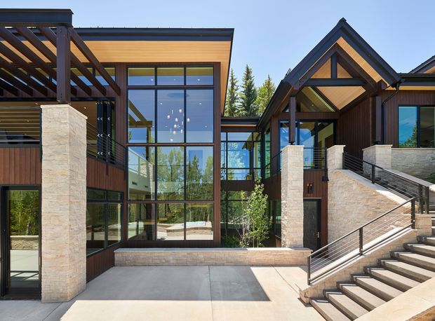 Exterior view of a contemporary multi-level residence with strong timber and dark metal detailing. The facade combines expansive floor-to-ceiling glazing, warm timber soffits and cladding, pale stone-clad structural columns, covered outdoor structures, and a broad stairway leading to the entry. A glazed connecting volume and surrounding trees are visible.