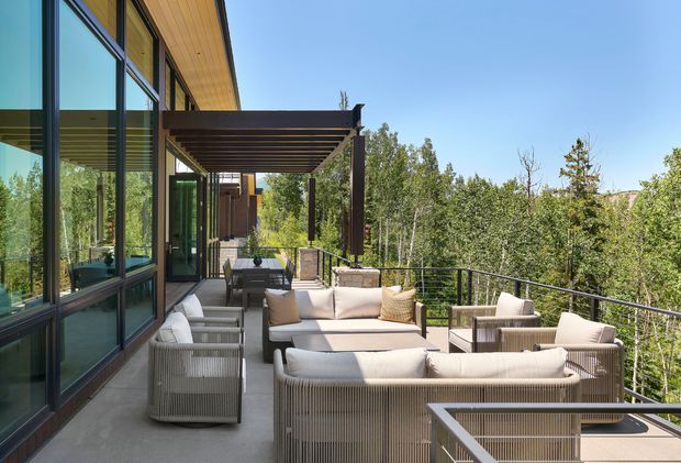 A spacious elevated outdoor terrace beside a contemporary glazed building, furnished with a large neutral-toned lounge setting and wicker-style armchairs. A dark timber pergola provides shade along the building, while black metal balustrading frames views into dense surrounding trees. The building features expansive floor-to-ceiling glazing, dark window frames, and warm timber soffit lining.