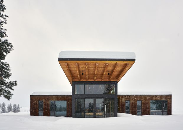 Front exterior view of a contemporary single-storey building in a snowy setting. The structure has dark timber-clad wings, extensive dark-framed glazing, and a dramatic projecting central entrance canopy with an exposed timber soffit and rounded light-coloured roof edge. Snow covers the ground and roof surfaces, while trees are visible at the sides.