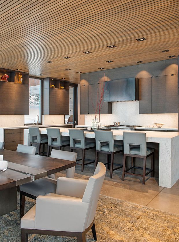 A sophisticated open-plan kitchen and dining area with a long island bench lined with upholstered bar stools, dark built-in cabinetry, a pale stone benchtop, integrated cooktop and rangehood, and warm timber slatted ceiling. The foreground contains a dark dining table and upholstered armchairs, while concealed and recessed lighting creates a refined, softly illuminated atmosphere.