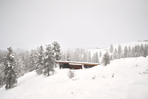 A low, elongated modern cabin or house sits partially embedded in a snowy alpine landscape. The building has a simple flat or very shallow-pitched roof with broad eaves, dark timber or metal wall surfaces, and a long glazed frontage. Snow-covered conifer trees and rolling mountain terrain surround the isolated structure, creating a quiet winter setting.