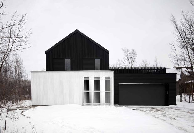 Exterior view of a compact contemporary house in a snowy, wooded setting. The building combines a tall black vertically ribbed gable-roof volume with a lower white vertically ribbed wing, a black garage door, and a screened white gate or privacy element. Bare deciduous trees and snow surround the property.