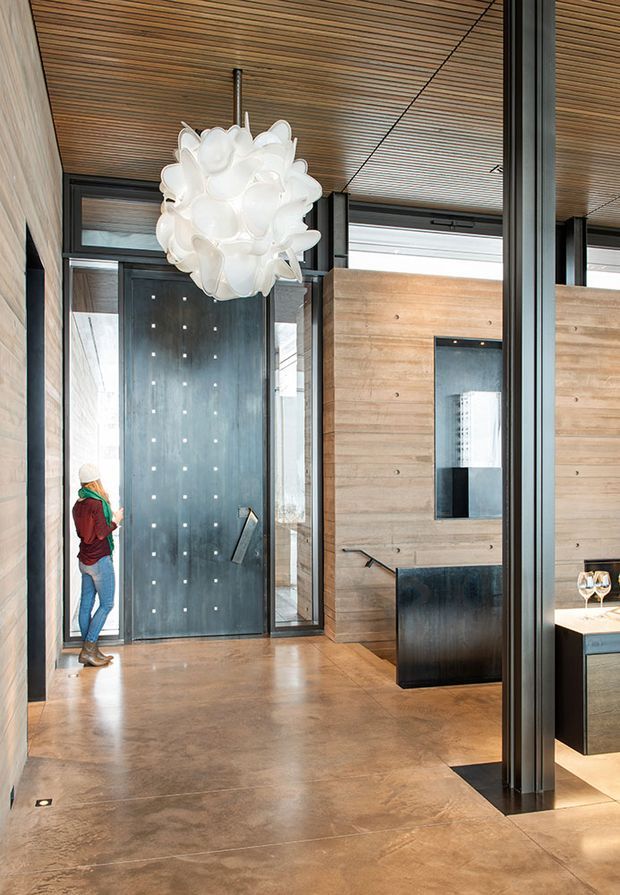 A sophisticated contemporary entry hall with a dramatic oversized sculptural pendant light, full-height glazing in dark metal frames, a tall dark pivot entrance door, warm timber slat ceiling, timber wall panelling, and polished concrete flooring. A partially visible kitchen or bar area appears beyond the partition on the right.