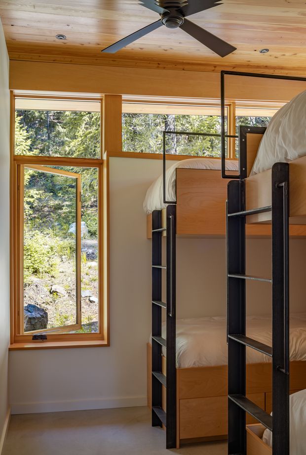 A compact bedroom with built-in bunk beds featuring warm timber bed frames and cabinetry, black metal ladder shelving and guard rails, and white bedding. The room has extensive timber ceiling lining, timber-framed windows, pale walls, and views of a wooded rocky landscape outside. A dark ceiling fan is centered overhead, contributing to a practical cabin-like feel.