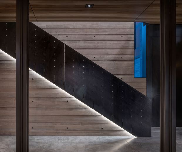 A dramatic contemporary stairway viewed through a dark-framed opening. The staircase has a solid black metal balustrade and concealed linear lighting beneath it, set against horizontal timber wall panelling. The ceiling continues the timber slat treatment, while a blue-toned glazed or illuminated vertical element adds contrast. The composition is minimalist, geometric, and moody.