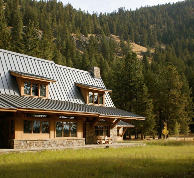 Exterior view of a rustic mountain lodge set in a forested, sloping landscape. The two-storey home has warm timber siding and trim, a grey standing-seam metal pitched roof with dormer windows, a stone-clad lower wall and chimney, and a covered timber veranda. A dense evergreen forest and rocky hillside form the backdrop.