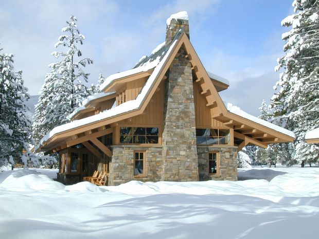 Snow-covered chalet-style house in a forested mountain setting, viewed from the front. The building has a steeply pitched, multi-gabled roof with broad overhanging eaves, exposed timber rafters and brackets, warm vertical timber cladding, and a prominent full-height natural stone chimney and facade feature. Large glazed openings sit beneath the roofline, with deep snow covering the surrounding ground.