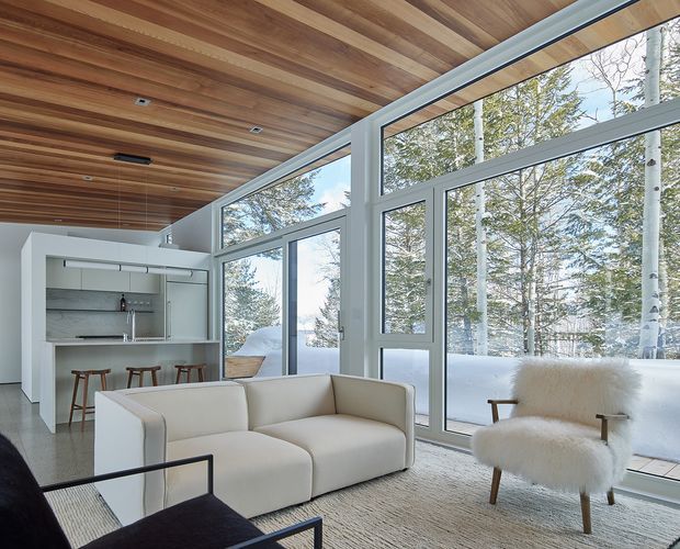 A bright, contemporary open-plan living room with a compact integrated bar or kitchenette, white upholstered seating, a black-framed lounge chair, and extensive floor-to-ceiling glazing overlooking a snowy wooded landscape. The room features a warm timber-lined sloped ceiling with recessed spotlights, pale walls, and a neutral textured rug.