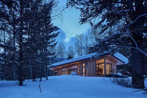 A low, modern cabin-like house set among dense snow-covered forest. The building has extensive floor-to-ceiling glazing, warm timber-clad walls, a broad gently sloping roof with deep overhangs, and warm interior lighting contrasting with the cool blue snowy landscape.