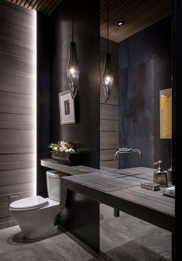 A dramatic contemporary bathroom with a wall-mounted vanity and integrated basin, large mirror, sculptural pendant lights, wall-mounted toilet, dark tiled and panelled walls, and a warm slatted timber ceiling. The space uses charcoal and grey finishes with metallic fixtures and moody accent lighting.