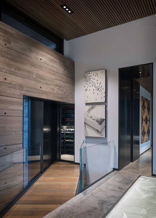 A sophisticated modern interior passage or landing with a dramatic concrete-look horizontal wall, dark timber slatted ceiling, black-framed glazing and doors, timber flooring, and a glass balustrade. A wine storage room is visible through the opening on the left, while framed monochrome artwork and a sculptural patterned wall feature add visual interest. The space appears to connect to a lower-level or open stair area.