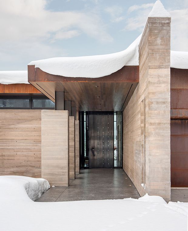 Exterior view of a contemporary, low-slung house entrance in a snowy mountain setting. The facade combines warm horizontal timber cladding, pale masonry or stone walls, large dark-framed glazing, a deep timber-lined recessed entry canopy, and a sculptural chimney. Heavy snow blankets the curved roof edges and foreground walkway.