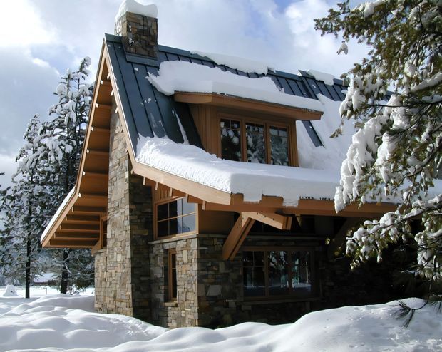 Snow-covered alpine house exterior with a steep pitched roof, dark metal roofing, heavy snow buildup, exposed timber eaves and balconies, and substantial natural stone chimney and wall cladding. The building is surrounded by snow-laden evergreen trees and appears designed for a cold mountainous setting.