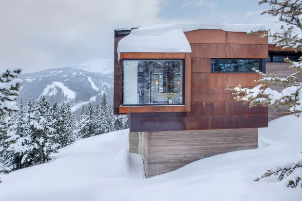 A compact contemporary mountain dwelling partially surrounded by deep snow, viewed from the exterior. The facade combines weathered reddish-brown timber cladding with a lighter horizontal timber base, large glazed windows, and a low, nearly flat roof heavily covered in snow. Snow-laden evergreen trees and a dramatic forested mountain slope form the background, emphasizing a remote alpine setting.