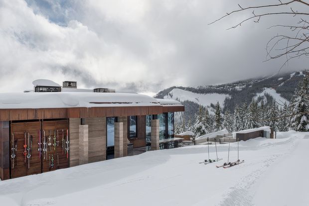 Snow-covered modern mountain home set in a heavily wooded alpine landscape. The low, flat-roofed building has warm timber cladding, large glazed openings, stone-like wall panels, and an outdoor ski storage area, with deep snow covering the roof and surrounding ground. Mountain slopes and snow-covered conifers form the background.