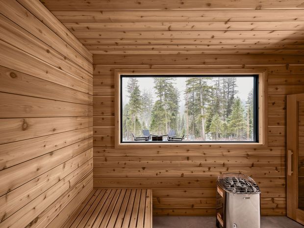 A compact wood-lined sauna with horizontal timber wall and ceiling boards, a built-in slatted timber bench, and a freestanding electric sauna heater. A large framed window looks onto a forested landscape, and a timber door is visible at right.