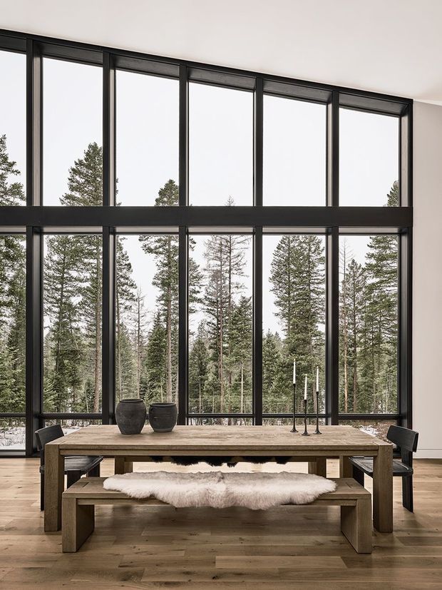 A refined dining area with a substantial rustic timber dining table and matching bench, positioned in front of expansive floor-to-ceiling black-framed glazing. Tall evergreen trees are visible outside, while black dining chairs, ceramic vessels, candleholders, and a sheepskin throw add restrained natural texture. The room has a very high ceiling and a calm, contemporary lodge-like character.