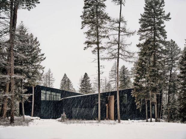 A low, angular modern house set within a dense snow-covered conifer forest. The building has dark, nearly black exterior cladding, extensive horizontal glazing on the left volume, and a tall warm timber-toned entrance or vertical feature panel near the centre. Snow blankets the foreground and roofline, creating a stark contrast with the dark geometric facade and surrounding trees.