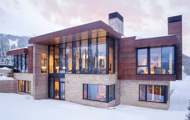 A contemporary multi-level mountain residence photographed in winter, with extensive floor-to-ceiling glazing, projecting flat roof forms, warm timber-clad soffits and upper walls, pale masonry lower walls, and dark framed windows. Snow-covered terrain and forested mountains are visible around the home, while warm interior lighting creates a welcoming evening atmosphere.