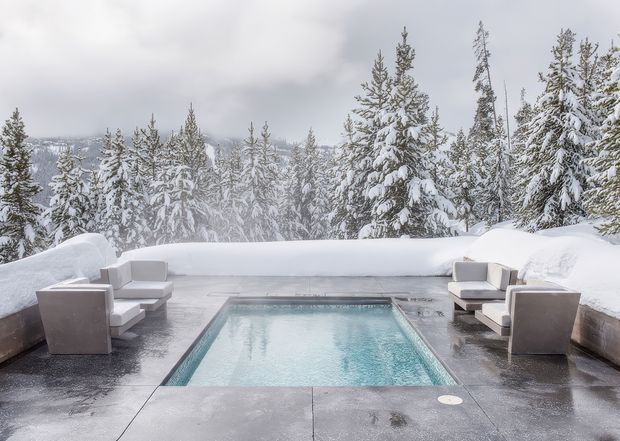 A rectangular outdoor pool is set into a dark paved terrace surrounded by deep snow. Minimalist cushioned lounge chairs line both sides, while snow-covered evergreen trees form a dense forest backdrop beneath an overcast sky. The scene has a luxurious, serene alpine-resort atmosphere.