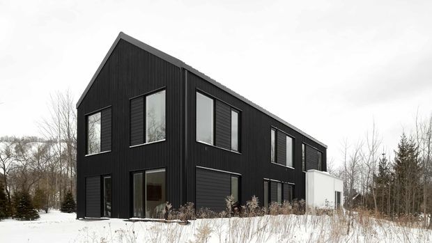 Exterior view of a contemporary two-storey black-clad house set in a snowy, wooded landscape. The building has a simple gable roof, vertically ribbed dark cladding, large rectangular windows, and a contrasting white rear extension.