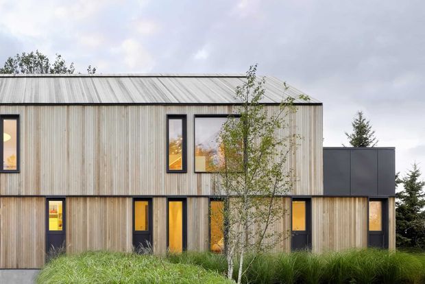 Exterior view of a contemporary two-storey house with a simple rectangular form, pale vertical timber cladding, dark window and door frames, and a low-pitched metal roof. Warmly lit windows punctuate the facade, while grasses and small trees form a landscaped foreground.
