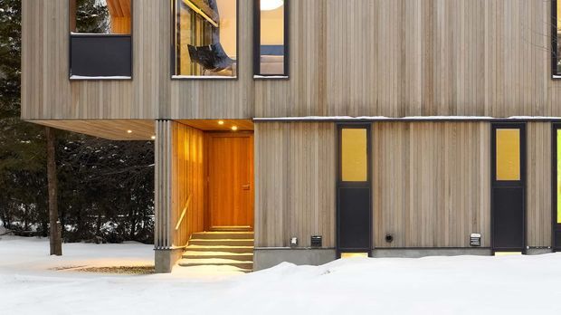 A modern multi-storey building exterior in a snowy, wooded setting. The facade combines vertical timber cladding with dark-framed windows and doors, warm timber-lined recessed entryways, and a projecting upper volume supported by a slender column. Snow covers the foreground, and the composition highlights the building envelope and entrances.