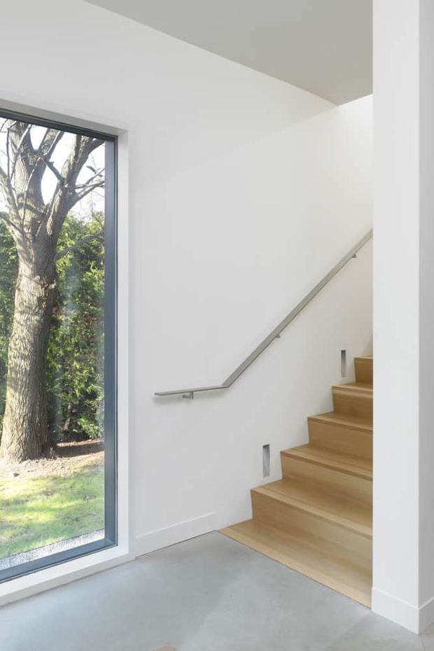 Bright minimalist interior stairway with a straight timber staircase, slim metal handrail, white walls and trim, and a large dark-framed window overlooking trees and lawn. The pale grey smooth floor and abundant natural light create a clean, contemporary feel.