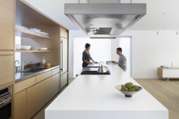 Bright open-plan contemporary kitchen viewed from behind a large white island. The kitchen features pale timber cabinetry, open shelving, integrated stainless-steel appliances, a sink, cooktop, kettle and a large stainless-steel rangehood. Two people sit at the island, with a sparse adjacent living area visible beyond. The space has white walls, light timber flooring and abundant daylight.