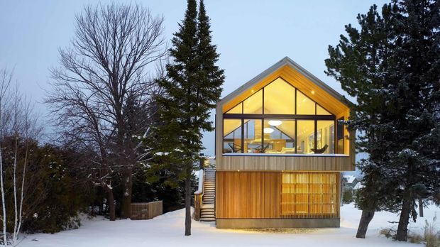 Exterior view of a compact contemporary timber-clad gable-roof house in a snowy, wooded setting. The upper level has a prominent glazed gable facade with warm interior lighting, while the lower level is clad in vertical timber boards with a screened section. Evergreen and deciduous trees frame the building.