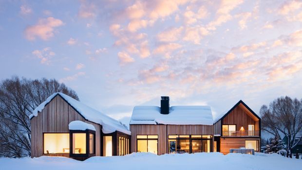 A contemporary timber-clad house in a snowy wooded setting, photographed at dusk. The low, elongated residence has multiple pitched gable roof volumes covered in snow, large illuminated windows, a central glazed living area, and a taller two-storey wing with a recessed balcony. Warm interior lighting contrasts with the cool blue winter landscape and pastel sky.