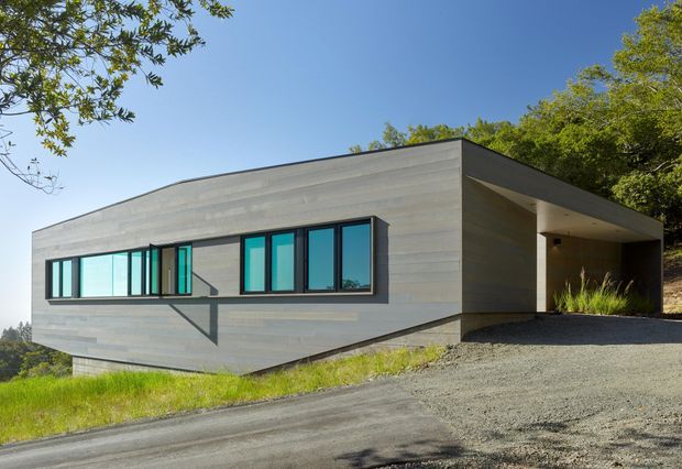 A low, rectilinear modern house set on a sloping, wooded site. The exterior is clad in horizontal grey concrete or cement-like panels, with a strong flat roof, dark-framed turquoise-tinted glazing, and a recessed covered entry or carport-like opening at the right. A gravel driveway and landscaped slope surround the building.