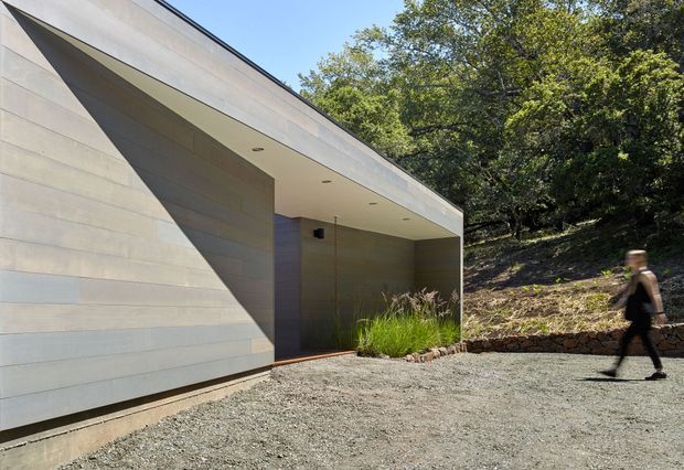 A minimalist modern building exterior with a sharply angled, low-profile roofline and an expansive recessed entry canopy. The facade is clad in pale horizontal panels, with a dark recessed doorway and discreet circular ceiling downlights. A gravel forecourt, grasses, and a low stone retaining wall connect the building to a wooded landscape. A blurred person provides scale.