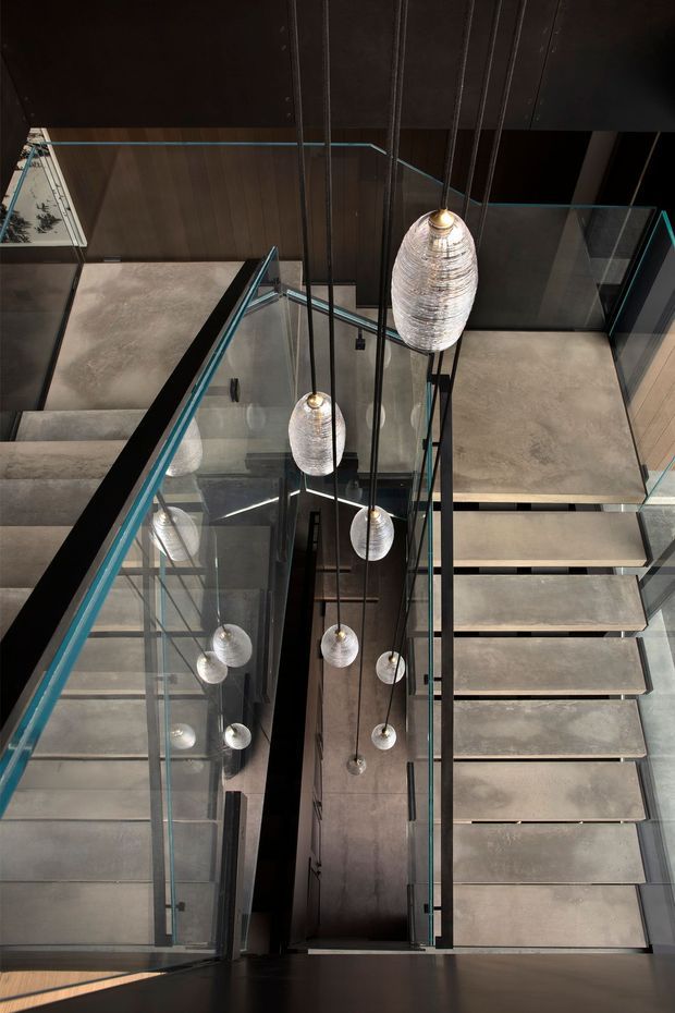 Looking down through a tall stairwell with a contemporary floating staircase, broad concrete or stone treads, dark metal stringers and handrails, and transparent glass balustrades. Multiple sculptural pendant lights hang through the stair void, with dark timber or painted surfaces framing the space. The composition emphasises vertical depth, reflections, and the double-height circulation void.