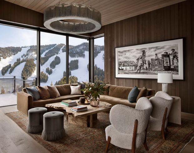 A warm, refined living room with expansive corner glazing overlooking a snow-covered mountainous landscape. The room features dark vertical timber wall panelling, a matching timber-lined ceiling, a large textured rug, a substantial sectional sofa, sculptural lounge chairs, ottomans, a low timber coffee table, and a dramatic circular pendant light. A framed black-and-white artwork and table lamp complete the layered, lodge-inspired interior.