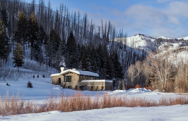 A secluded single-storey house set in a snowy alpine landscape. The home has a low, gently sloping roof, timber-clad walls, large dark-framed windows, and a masonry or stone chimney emitting smoke. Snow-covered conifer forest and rugged mountain slopes surround the property, with dormant grasses and shrubs in the foreground.
