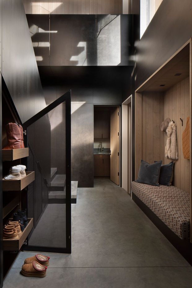 A narrow, double-height residential entry hall with a dark, industrial-inspired fit-out. The space features a polished grey concrete floor, dark metal and black mesh screening, charcoal wall panels, timber wall panelling and built-in joinery. A built-in upholstered bench with cushions and coat hooks occupies the right side, while open shelving and concealed storage line the left. Strong sunlight enters from high-level glazing, creating dramatic geometric shadows.