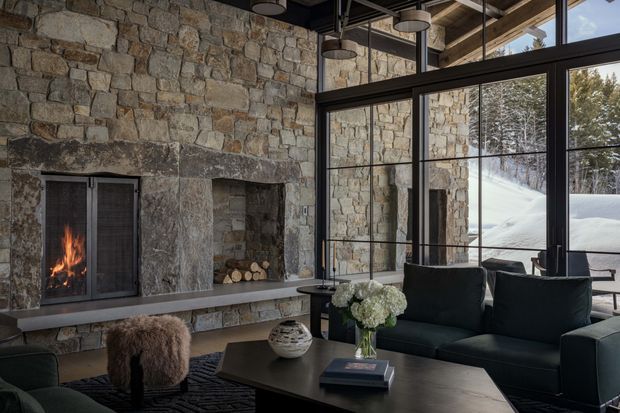 A cozy, high-end living room in a stone-clad mountain setting. The room features a large rustic stone fireplace with a lit fire and log storage, expansive black-framed floor-to-ceiling glazing, exposed timber ceiling structure, and dark contemporary sofas arranged around a substantial dark coffee table. Snow-covered trees and terrain are visible outside.