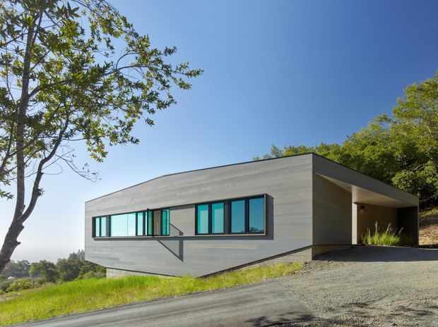 A low, single-storey modern house set on a sloping, leafy site. The building has a strong horizontal form, flat roof, extensive dark-framed glazing, and smooth grey horizontal cladding. A recessed covered entry or carport-like opening is visible on the right, while a gravel driveway curves across the foreground. Trees and distant landscape surround the minimalist residence.