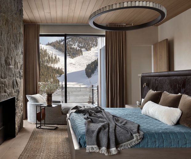 A refined mountain bedroom with a large bed facing floor-to-ceiling glazing and a snowy, forested alpine landscape beyond. The room features a textured stone fireplace wall, warm timber plank ceiling, dark framed glass doors, full-height brown curtains, a sculptural circular pendant light, and layered neutral bedding with a blue quilt and grey throw. A small seating area and patterned rug add comfort and warmth.